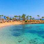 Tropical beach with crystal-clear turquoise water, white sand, palm trees, and beach loungers under a blue sky.