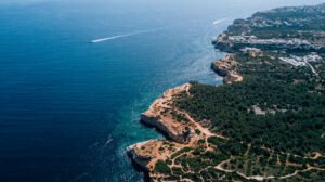 Aerial view of a rocky coastline with turquoise waters, greenery, and a speedboat leaving a trail.
