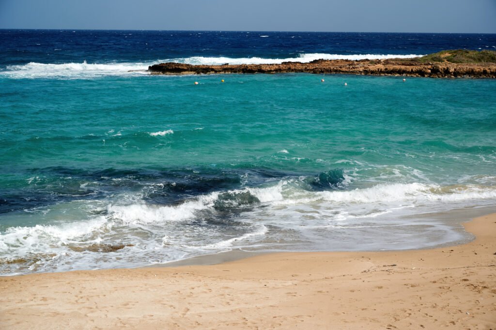 Beautiful turquoise ocean waves crashing on a sandy beach with clear blue water and rocky outcrop in the distance.