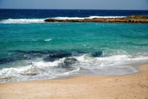 Beautiful turquoise ocean waves crashing on a sandy beach with clear blue water and rocky outcrop in the distance.