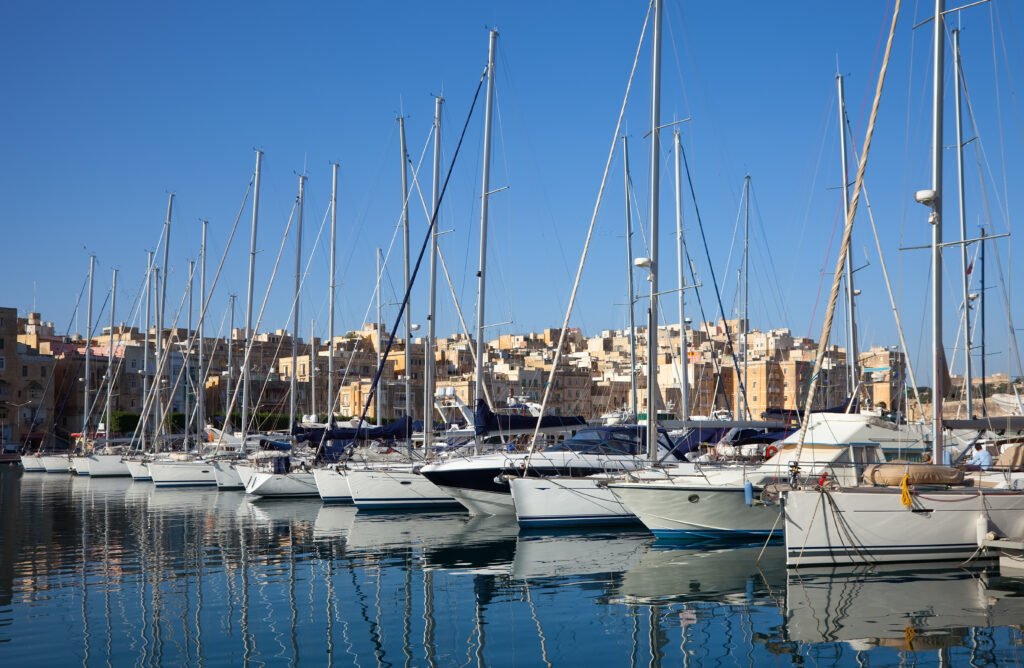 Row of sailboats docked in a calm marina with clear blue water and city buildings in the background
