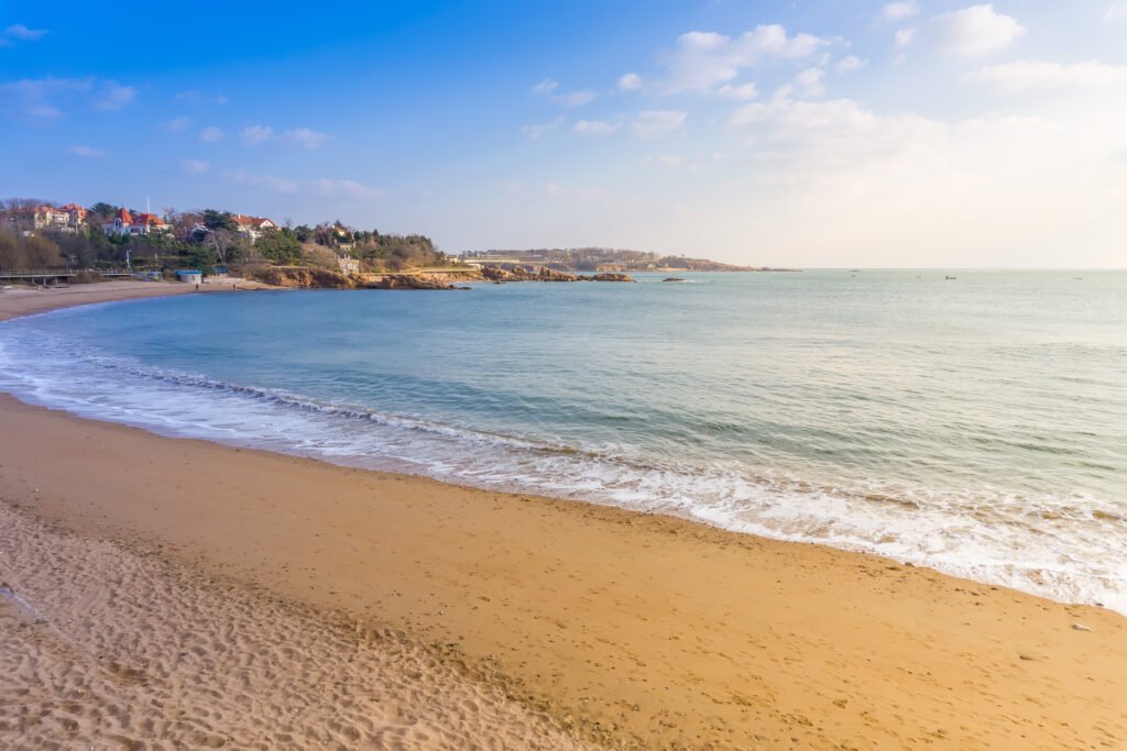 Sandy beach with gentle waves under a clear blue sky, lined with coastal buildings and rocky outcrops in the distance.